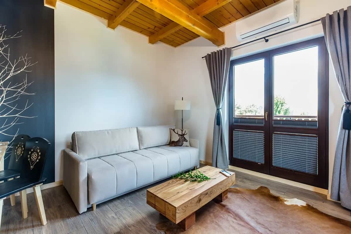 Salon z jasną sofą i wyjściem na balkon z roletami