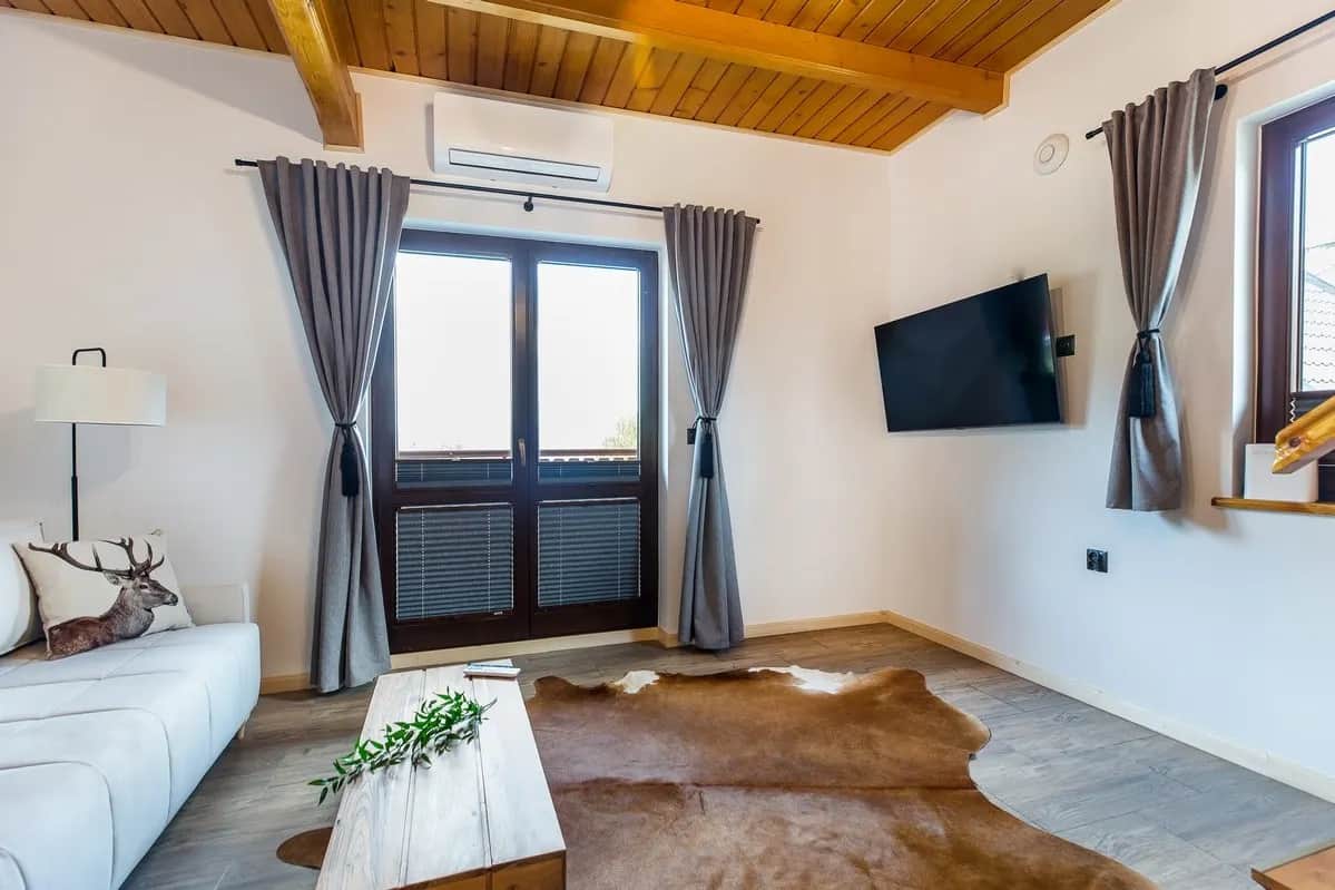 Jasny salon z białą sofą, drewnianym sufitem, telewizorem na ścianie i przeszklonymi drzwiami na balkon, na podłodze skóra bydlęca