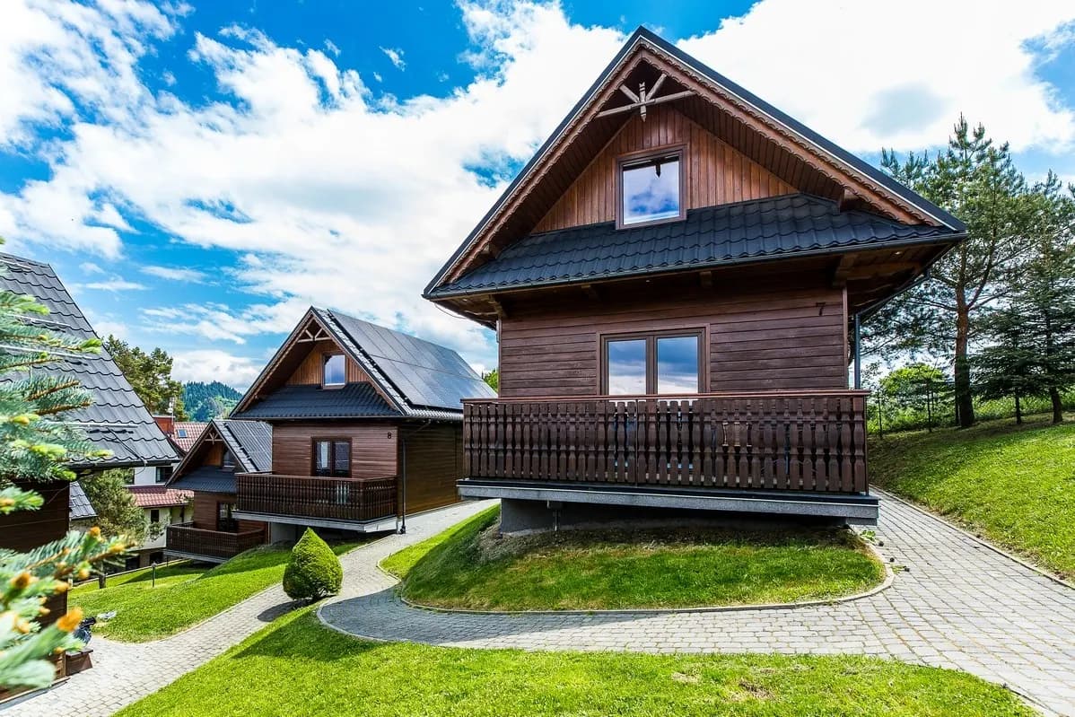 Drewniany domek z dwuspadowym dachem i balkonem, widziany z poziomu ogrodu, w tle drugi domek i zielone wzgórza