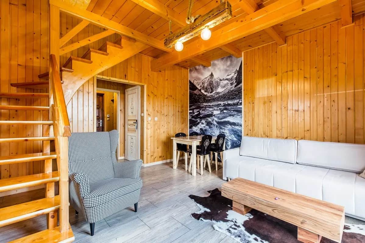 Salon z drewnianymi schodami na antresolę, fotelem w pepitkę i kącikiem jadalnym przy fototapecie górskiej