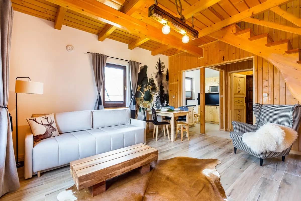 Jasny salon z drewnianym stropem belkowym, szarą kanapą, drewnianym stolikiem, skórą bydlęcą na podłodze i otwartym przejściem do jadalni i kuchni