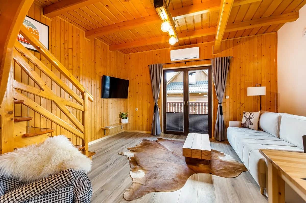 Salon z drewnianą boazerią, telewizorem na ścianie, kanapą, drewnianymi schodami na poddasze i przeszklonymi drzwiami na balkon